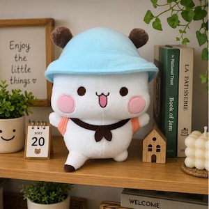 May include: A white plush toy featuring a light blue bucket hat, brown ears, and pink cheeks. The character has a happy expression with a tongue sticking out, a dark brown neckerchief, and orange backpack straps visible on its shoulders.