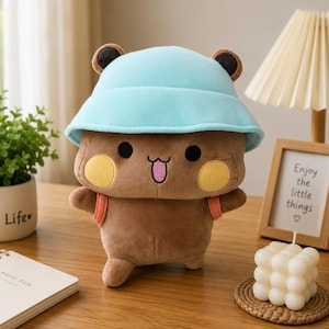 May include: A brown plush toy featuring a light blue bucket hat, round yellow cheeks, black eyes, and a small pink tongue. The stuffed animal has small dark brown ears and orange straps on its sides, resembling a small backpack or accessory.