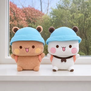 May include: Two plush toys featuring round heads and bodies, wearing light blue bucket hats. One toy is brown with yellow cheeks, the other is white with pink cheeks and a dark brown neck scarf. Both have small ears and peach-colored backpacks.