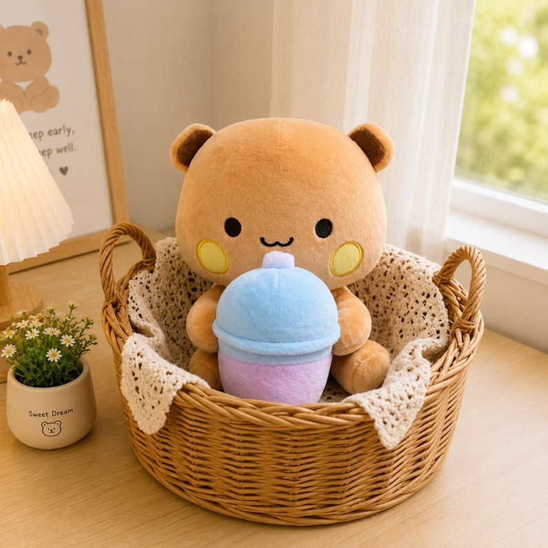 May include: A light brown plush bear toy with round ears, black eyes, and yellow circular cheeks. The bear is holding a small, two-toned blue and purple plush drink cup. The toy is sitting inside a round, woven brown wicker basket.