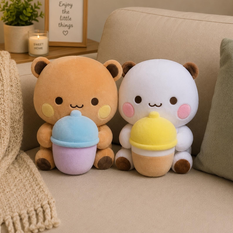 May include: Two plush bear toys sitting side by side. The left bear is light brown with yellow cheeks, holding a blue and lavender cup. The right bear is white with pink cheeks, holding a yellow and tan cup. Both have dark brown ears and paws.