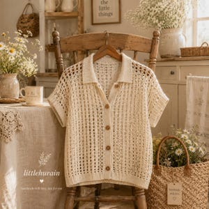 May include: A cream-colored, short-sleeved, button-down knit shirt featuring an open-weave, grid-like pattern, a classic collar, and five visible buttons. The garment is displayed on a wooden hanger against a rustic, light-filled interior.