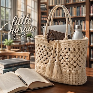 May include: A cream-colored crochet tote bag featuring an openwork diamond pattern, two sturdy handles, and two decorative tassels hanging from the front. The bag contains a tablet, a dark notebook, glasses, and a silver water bottle.