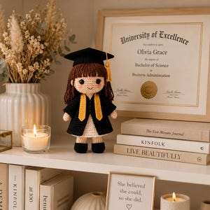 May include: A crocheted doll wearing a black graduation cap with a yellow tassel, a black graduation gown, and a white dress. The doll has brown hair, rosy cheeks, and black button eyes. It stands upright, featuring a detailed yarn construction.