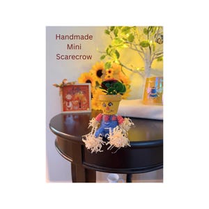 May include: A handmade mini scarecrow figurine featuring a yellow painted terracotta pot head with a drawn face, wearing blue overalls over a pink plaid shirt, with raffia straw accents at the wrists and ankles, topped with a small hat.