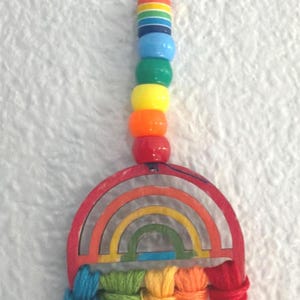 Peut inclure: Une décoration suspendue composée d'un arc-en-ciel en bois peint en rouge, orange, jaune, vert et bleu. Sous l'arc se trouvent cinq pompons en fil de laine bleu, vert, jaune, orange et rouge. Un cordon bleu foncé retient des perles multicolores.
