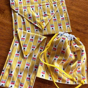May include: Two fabric envelopes and one drawstring pouch featuring a repeating yellow pencil pattern with black tips, grey ferrules, and pink erasers. The envelopes have triangular flaps with yellow closures, and the pouch has yellow drawstrings.
