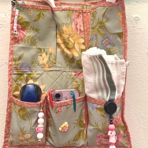 May include: A fabric wall organizer featuring a sage green background with a floral pattern of pink roses and yellow sunflowers. It has multiple pockets containing a white brush, a green duster, a white towel, pens, and decorative beaded charms.