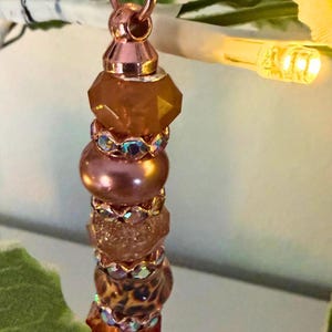 May include: A vertical decorative ornament featuring a series of stacked beads in shades of amber, copper, and brown. The beads are separated by small, metallic spacers with iridescent crystals and are suspended from a light peach ribbon loop.