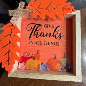 May include: A square wooden shadow box frame featuring an orange background with the black text GIVE THANKS IN ALL THINGS. The frame is decorated with orange felt leaves and a jute twine bow, with colorful pumpkin and leaf cutouts at the base.