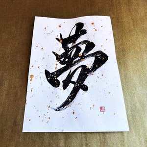 以下が含まれることがあります： 白い紙に黒いインクで書かれた書道。金色の飛沫が散りばめられている。書道は「夢」という漢字で、夢を意味する。