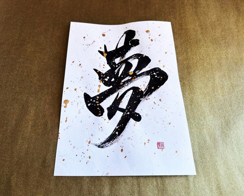 以下が含まれることがあります： 白い紙に黒いインクで書かれた書道。金色の飛沫が散りばめられている。書道は「夢」という漢字で、夢を意味する。