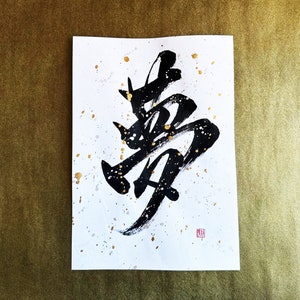 以下が含まれることがあります： 白い紙に黒いインクで書かれた書道。金色の絵の具が飛び散っている。書道は「勇」という漢字で、「勇気」を意味する。