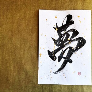 以下が含まれることがあります： 白い紙に黒いインクで書かれた書道。金色の絵の具が飛び散っている。書道は「夢」という漢字で、夢という意味です。