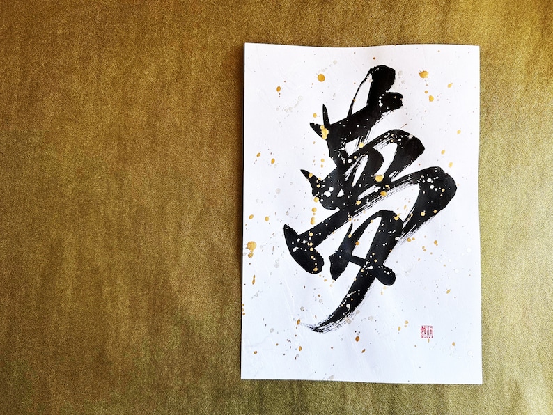 以下が含まれることがあります： 白い紙に黒いインクで書かれた書道。金色の絵の具が飛び散っている。書道は「夢」という漢字で、夢という意味です。