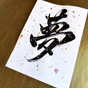 以下が含まれることがあります： 白い紙に黒いインクで書かれた書道作品。金色の飛沫が散りばめられている。書道は「夢」という漢字で、夢を意味する。