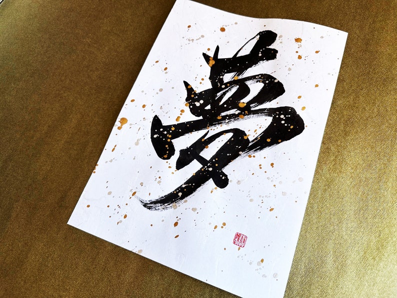 以下が含まれることがあります： 白い紙に黒いインクで書かれた書道作品。金色の飛沫が散りばめられている。書道は「夢」という漢字で、夢を意味する。