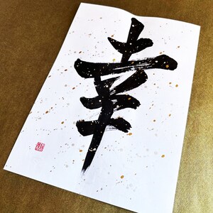 May include: A black ink calligraphy piece on white paper with gold and white splatters. The calligraphy depicts the Japanese character for "happiness".