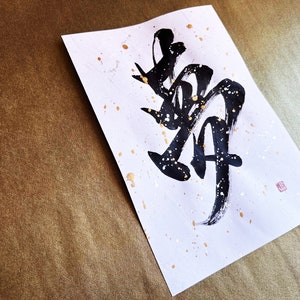 以下が含まれることがあります： 白い紙に黒い墨で書かれた日本の文字「舞」（「まい」と読む）は「ダンス」を意味します。紙には金色の斑点があり、右下に小さな赤い印があります。