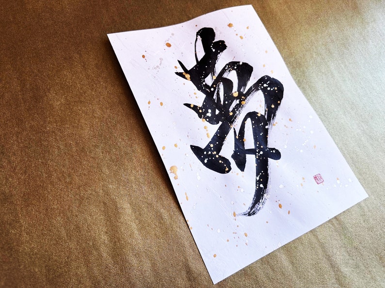 以下が含まれることがあります： 白い紙に黒い墨で書かれた日本の文字「舞」（「まい」と読む）は「ダンス」を意味します。紙には金色の斑点があり、右下に小さな赤い印があります。