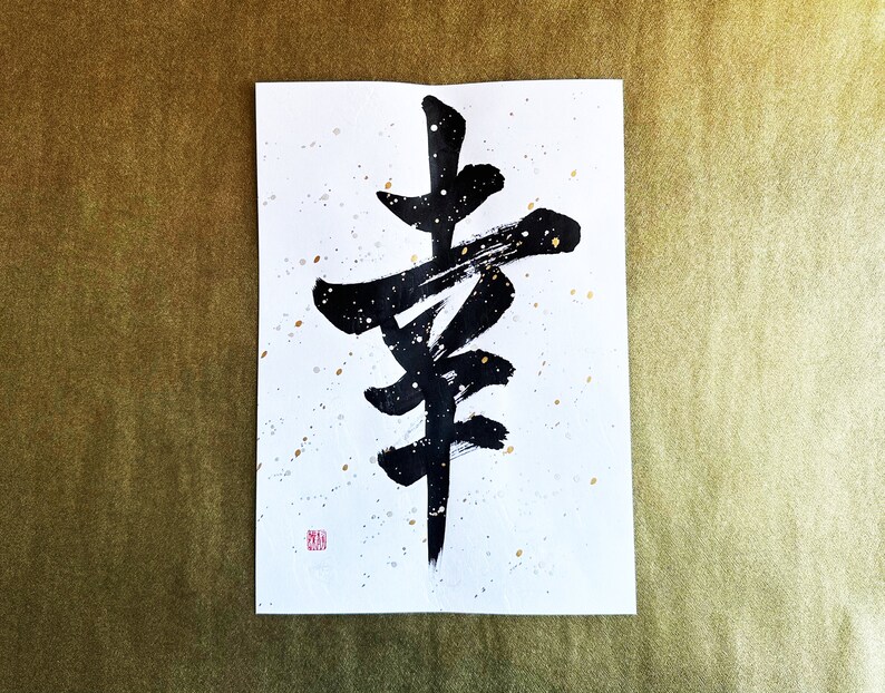 May include: Black ink calligraphy on white paper with gold speckles. The calligraphy depicts the Japanese character for "happiness".