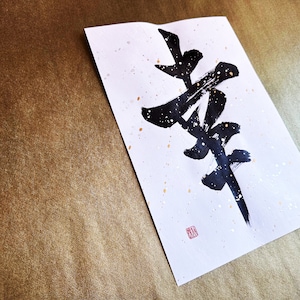 May include: A white paper with black calligraphy writing on it. The calligraphy is of the Japanese character "Zen". The paper has gold flecks on it and a red seal in the bottom right corner.