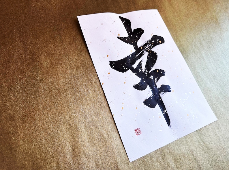 May include: A white paper with black calligraphy writing on it. The calligraphy is of the Japanese character "Zen". The paper has gold flecks on it and a red seal in the bottom right corner.