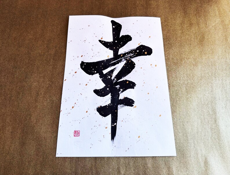 May include: Black ink calligraphy on white paper with gold speckles. The calligraphy depicts the Japanese character for "happiness".