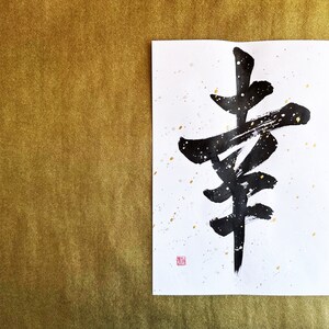 May include: Black ink calligraphy on white paper with gold accents. The calligraphy depicts the Japanese character for "happiness".
