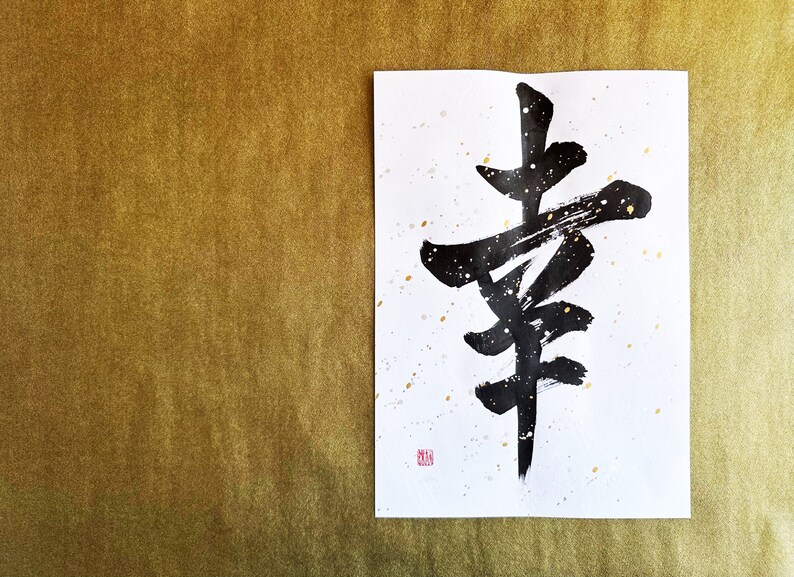 May include: Black ink calligraphy on white paper with gold accents. The calligraphy depicts the Japanese character for "happiness".