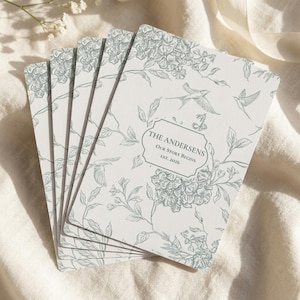Peut inclure: Une pile de cartes rectangulaires blanches avec un motif botanique au trait vert sauge représentant des fleurs et des oiseaux. Le centre de la carte supérieure présente un cadre décoratif avec le texte THE ANDERSENS, OUR STORY BEGINS, EST. 2026.