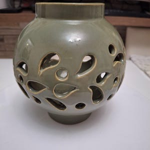 Könnte beinhalten: Eine kugelförmige Keramikvase mit glänzender salbeigrüner Glasur. Der Körper der Vase weist zahlreiche tropfenförmige Ausschnitte auf, die in einem verstreuten Muster über die Oberfläche verteilt sind und das hohle Innere durch das offene Design enthüllen.