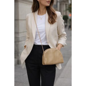 May include: A beige blazer worn over a white crew neck t-shirt and black trousers, paired with a tan semi-circular crossbody bag featuring a thin shoulder strap. The outfit is completed with a delicate gold necklace with a small pendant.