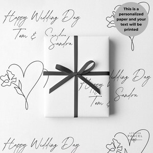 Op de afbeelding: Een rechthoekige geschenkdoos verpakt in wit papier met zwarte lijntekeningen van harten, bloemen en de tekst Happy Wedding Day Tom & Sandra. Een zwart lint is in het midden tot een strik gebonden. Een grijze ronde badge geeft aan dat dit papier gepersonaliseerd wordt.