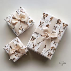 Pode incluir: Três caixas de presente retangulares embrulhadas em papel branco com um padrão de aguarela repetitivo de várias raças de cães e o texto Best dog mom. Cada caixa está atada com uma fita de cetim bege larga e lisa num grande laço.
