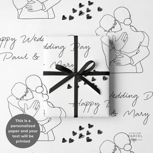 Può includere: Carta da regalo bianca con un'illustrazione a linee nere di una coppia che si abbraccia, cuori neri sparsi e il testo ripetuto Happy Wedding Day Paul & Mary, legata con un nastro nero. La carta include il testo Atelier Parcel.