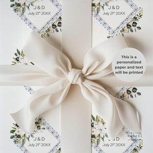 Könnte beinhalten: Weißes Geschenkpapier mit einem sich wiederholenden Rautenmuster, blauen Blumenrändern und grünem Blattwerk. Ein breites, cremefarbenes Satinband ist mittig zu einer großen Schleife gebunden. Der Text lautet J & D July 21st 20XX und This is a personalized paper.