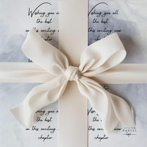 Op de afbeelding: Een witte geschenkdoos verpakt met een breed crèmekleurig satijnen lint gestrikt in een grote strik. De doos heeft een lichtgrijs marmerpatroon en zwarte tekst met de tekst, Wishing you all the best on this exciting new chapter, Atelier Parcel.