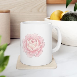 Peut inclure: Un mug en céramique blanche orné d'une illustration centrée d'une pivoine rose tendre aux pétales superposés. Le mug présente un corps cylindrique lisse et une anse incurvée sur le côté droit, posé sur un dessous de verre carré.