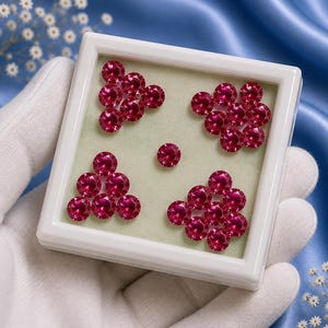 May include: A square white display case containing twenty-five small, round, faceted, deep pink gemstones arranged in four triangular clusters with one single gemstone placed in the centre of the light green velvet-textured insert.