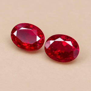 May include: Two oval-cut gemstones with a deep, vibrant red colour. Each stone features a faceted surface that reflects light, creating a brilliant, sparkling appearance. The gems are positioned side-by-side, showcasing their symmetrical shape.