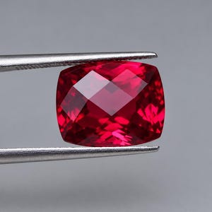 Può includere: Una gemma rossa taglio a cuscino tenuta da una pinzetta in metallo argentato. La pietra presenta una superficie sfaccettata che riflette la luce, creando riflessi cremisi profondi e scarlatti sulla sua forma quadrata lucida.