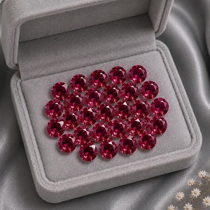 Könnte beinhalten: Eine Ansammlung von runden, facettierten, tiefroten Rubinen, die in einer mit grauem Samt ausgekleideten Schmuckschatulle angeordnet sind. Die Edelsteine weisen eine brillante, reflektierende Oberfläche mit zahlreichen Facetten auf.