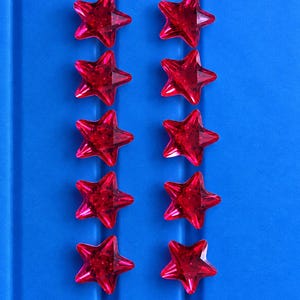 Peut inclure: Dix petites décorations en forme d'étoile à cinq branches, fabriquées dans un matériau rouge profond translucide. Chaque étoile présente une surface à facettes qui capte la lumière, disposée en deux colonnes verticales sur un fond bleu vif.
