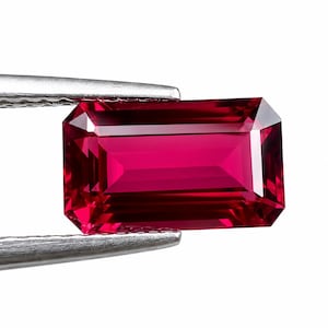 May include: A vibrant, deep red gemstone with a rectangular emerald cut, featuring sharp, clean facets that reflect light. The stone is held by silver-toned metal tweezers, showcasing its transparent, saturated color and precise geometric shape.