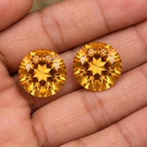 Puede incluir: Dos piedras preciosas de citrino redondas y facetadas con un tono amarillo dorado vibrante. Cada piedra presenta un corte preciso en forma de estrella en el centro, reflejando la luz con gran claridad y brillo a través de sus múltiples facetas.