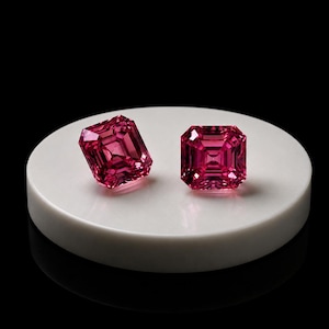 May include: Two vibrant, deep pink, square-cut gemstones with faceted surfaces. The stones are positioned side-by-side on a smooth, circular, white platform. The faceted cuts create intricate geometric patterns that reflect light across the surface.