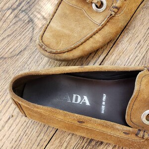 May include: A pair of brown suede loafers with silver buckle accents. The loafers have a leather sole and are made in Italy. The inside of one loafer shows the Prada logo and "Made in Italy" text.