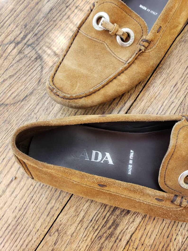 May include: A pair of brown suede loafers with silver buckle accents. The loafers have a leather sole and are made in Italy. The inside of one loafer shows the Prada logo and "Made in Italy" text.