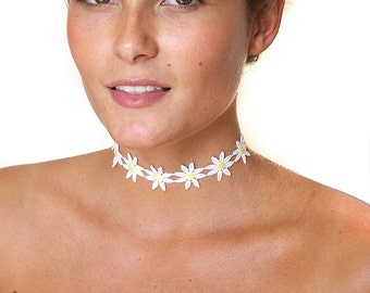 May include: A white daisy flower choker necklace with yellow centres. The necklace is worn by a person with brown hair against a white background.
